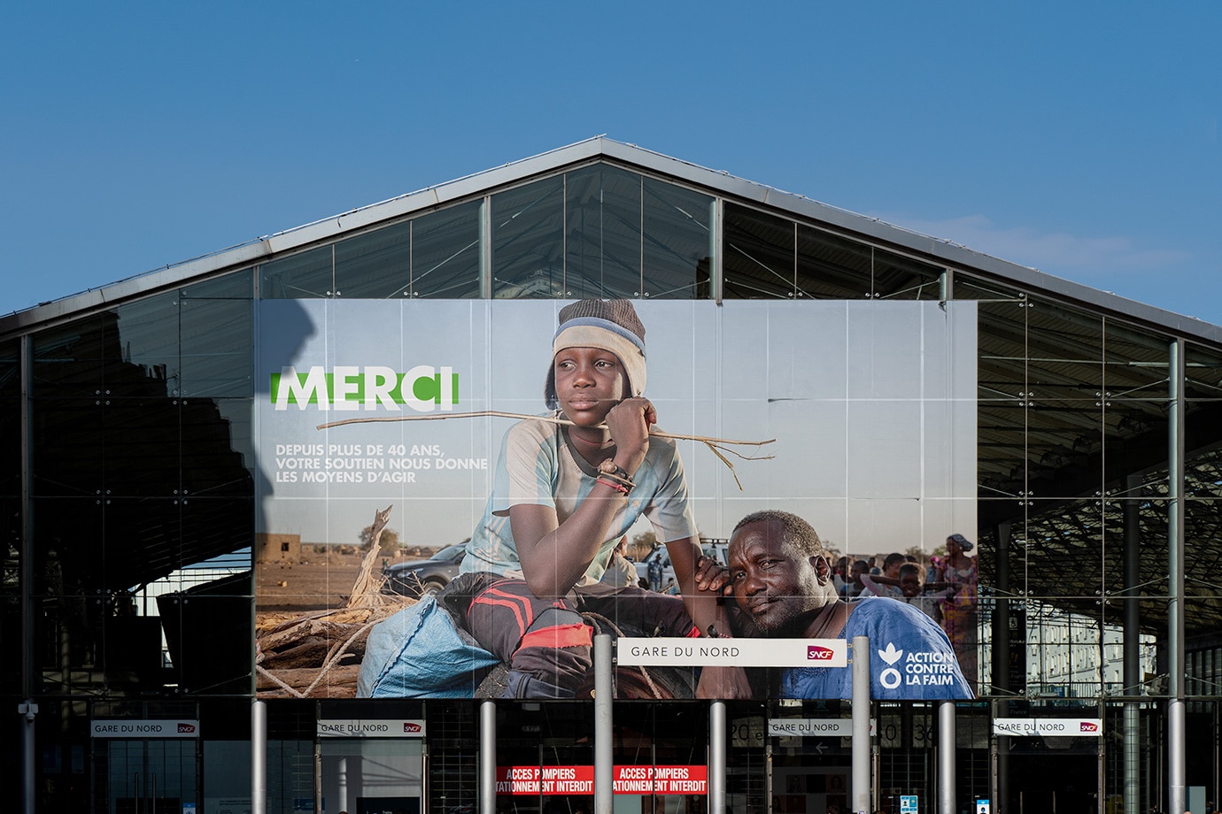Grande affiche publicitaire sur la façade extérieure de la Gare du Nord. L'affiche présente un jeune garçon pensivement assis, regardant au loin, à côté de son père souriant, avec le texte 'MERCI - Depuis plus de 40 ans, votre soutien nous donne les moyens d'agir' en grandes lettres, soulignant l'anniversaire de l'ONG Action contre la Faim.