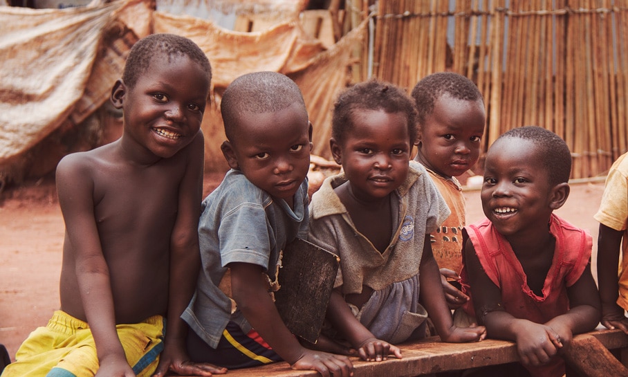 Enfants centrafricains souriants assis sur un banc dans un camp de réfugiés, avec des abris de fortune en arrière-plan, symbolisant la résilience et l'espérance dans des conditions de vie difficiles.