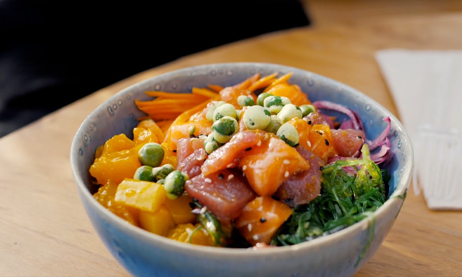 Bol de poke coloré avec des morceaux de poisson cru, des légumes frais, et des pois verts croquants, servi dans un bol en céramique bleue sur une table en bois.