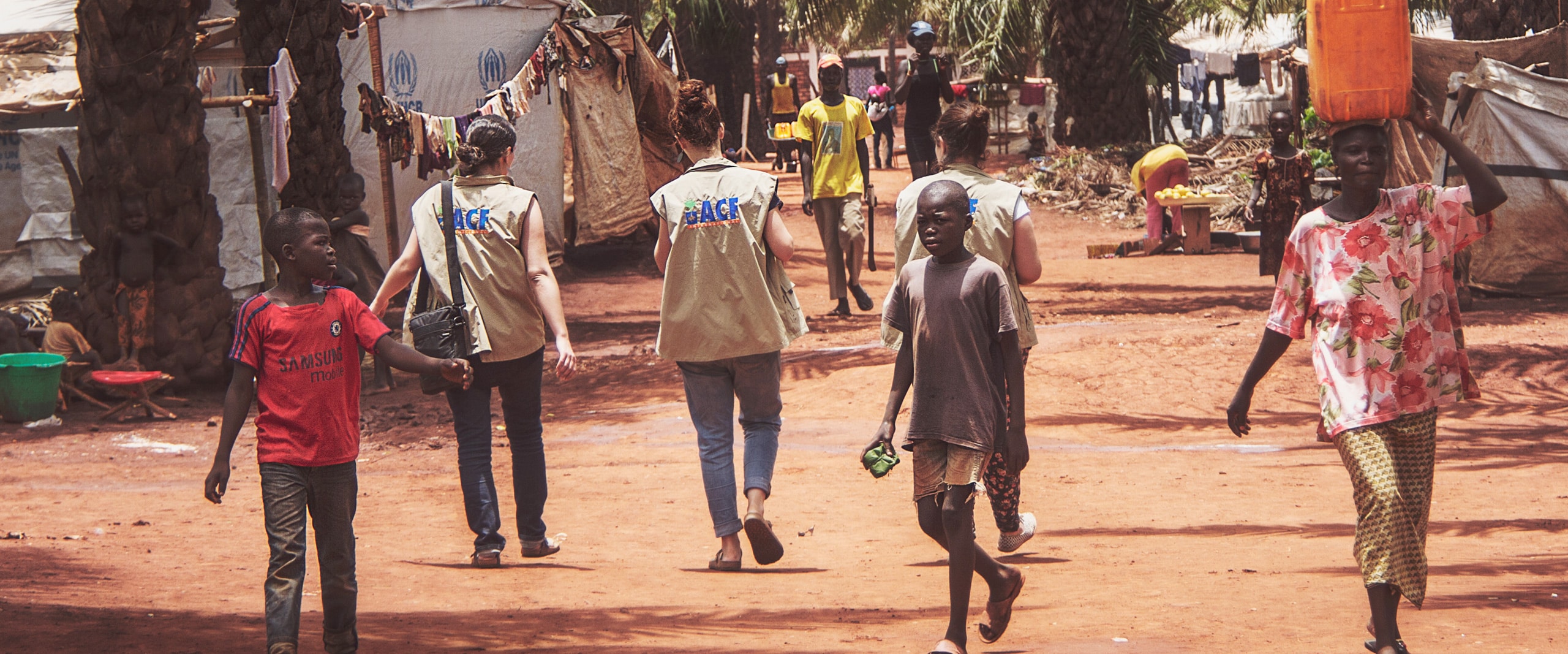 Deux humanitaires vêtus de gilets avec le logo 'ACF' (Action Contre la Faim) marchent dans un camp de réfugiés à Bangui en République centrafricaine, suivis par des enfants locaux. Les tentes de l'UNHCR sont visibles en arrière-plan, érigées dans un environnement de terre rouge et sous un ciel clair. L'image capture un moment de la vie quotidienne dans le camp, soulignant l'interaction entre les travailleurs humanitaires et la communauté.