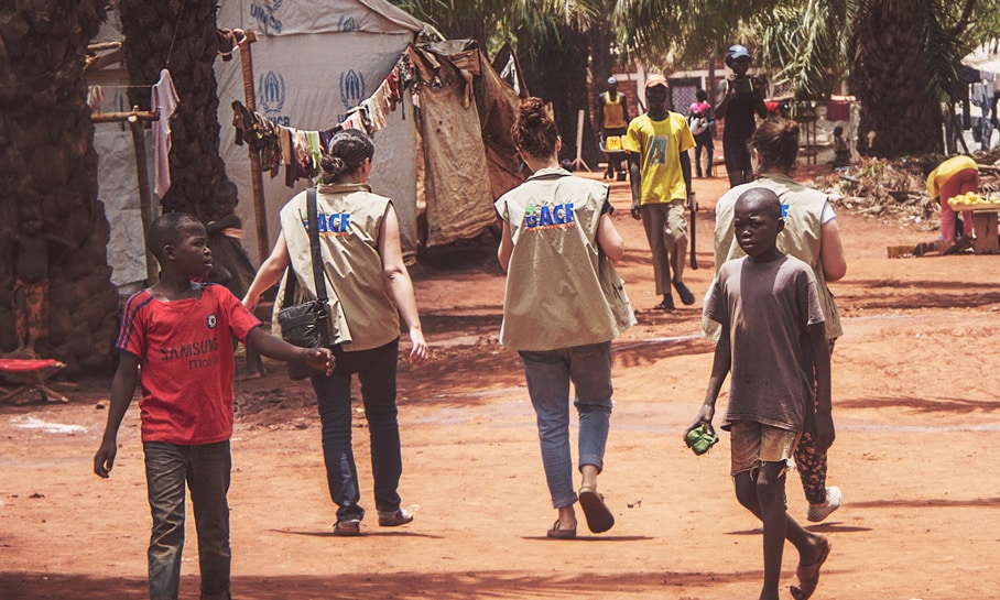 Deux humanitaires vêtus de gilets avec le logo 'ACF' (Action Contre la Faim) marchent dans un camp de réfugiés à Bangui en République centrafricaine, suivis par des enfants locaux. Les tentes de l'UNHCR sont visibles en arrière-plan, érigées dans un environnement de terre rouge et sous un ciel clair. L'image capture un moment de la vie quotidienne dans le camp, soulignant l'interaction entre les travailleurs humanitaires et la communauté.