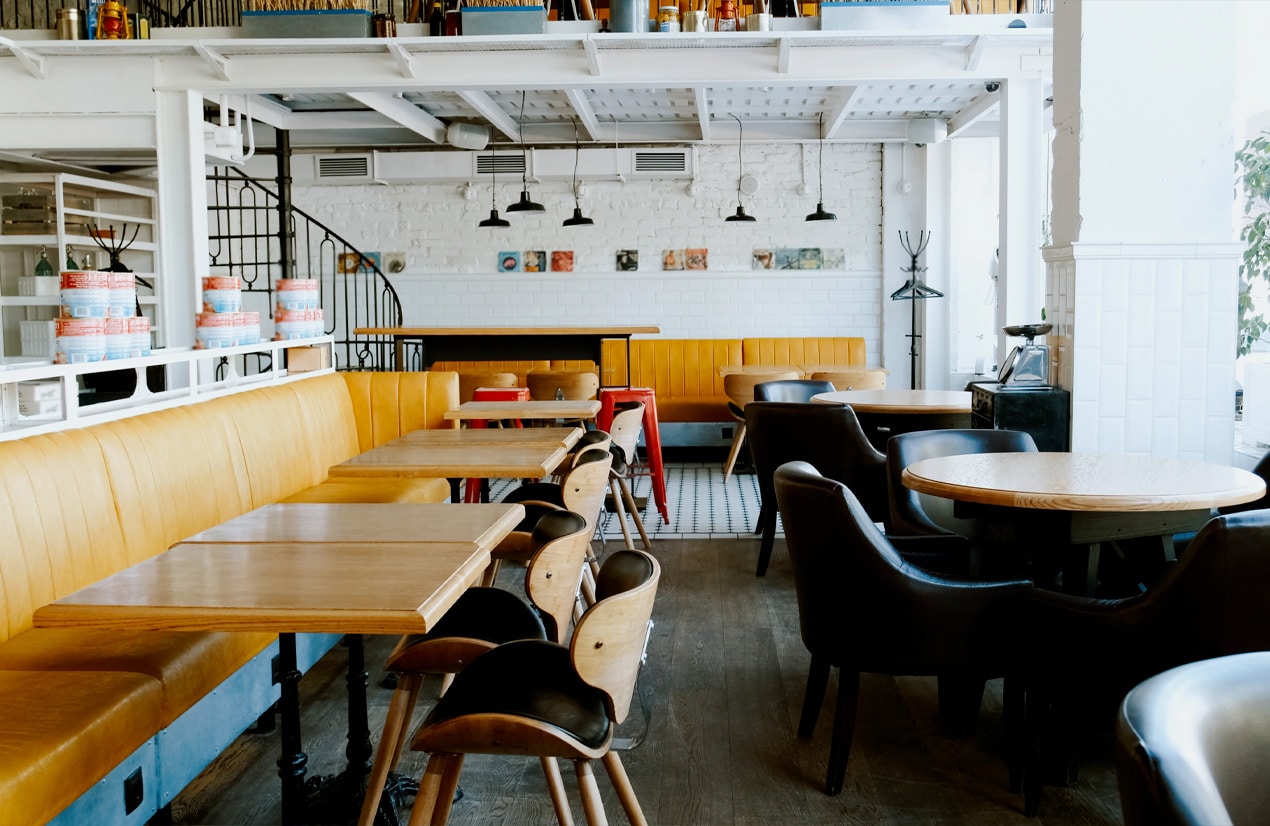 Intérieur d'un café moderne, vide, avec des sièges en cuir marron et des bancs en cuir jaune, des tables en bois, un sol en parquet et carrelage, et une décoration épurée avec des étagères blanches et des luminaires suspendus.
