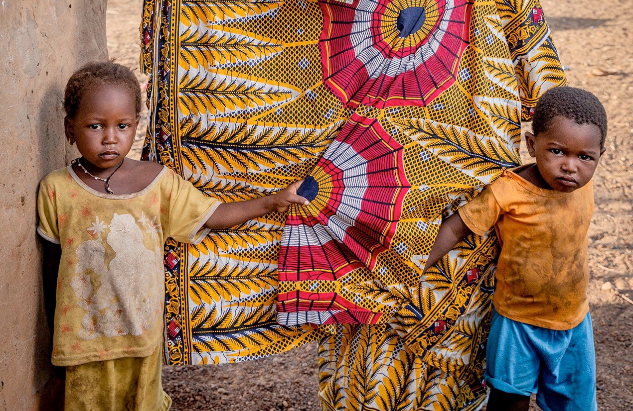 Deux jeunes enfants africains se tenant aux vêtements traditionnels colorés d'un adulte, symbolisant la protection et la culture en Afrique.
