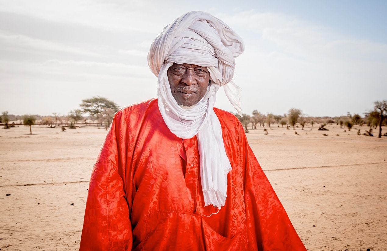 Homme africain vêtu de vêtements traditionnels rouges et blancs, portant un turban, se tenant dans un paysage désertique.
