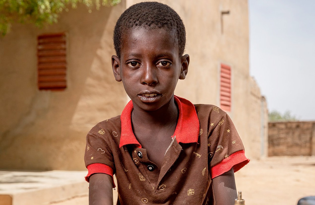 Un garçon portant une chemise brune avec un col rouge se tient devant une maison en terre cuite avec des volets rouges.