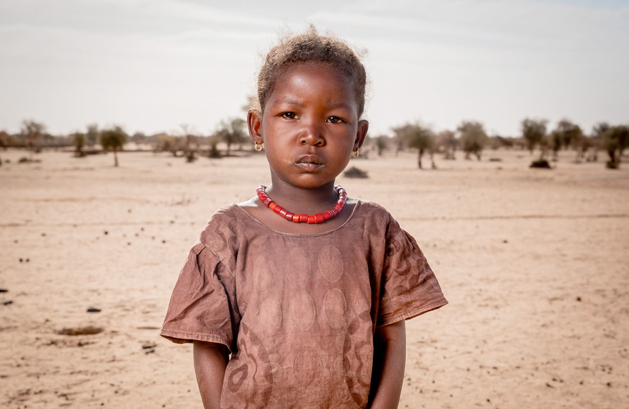 Une petite fille africaine portant un collier rouge et une robe marron se tient debout dans un paysage désertique avec des arbres en arrière-plan.