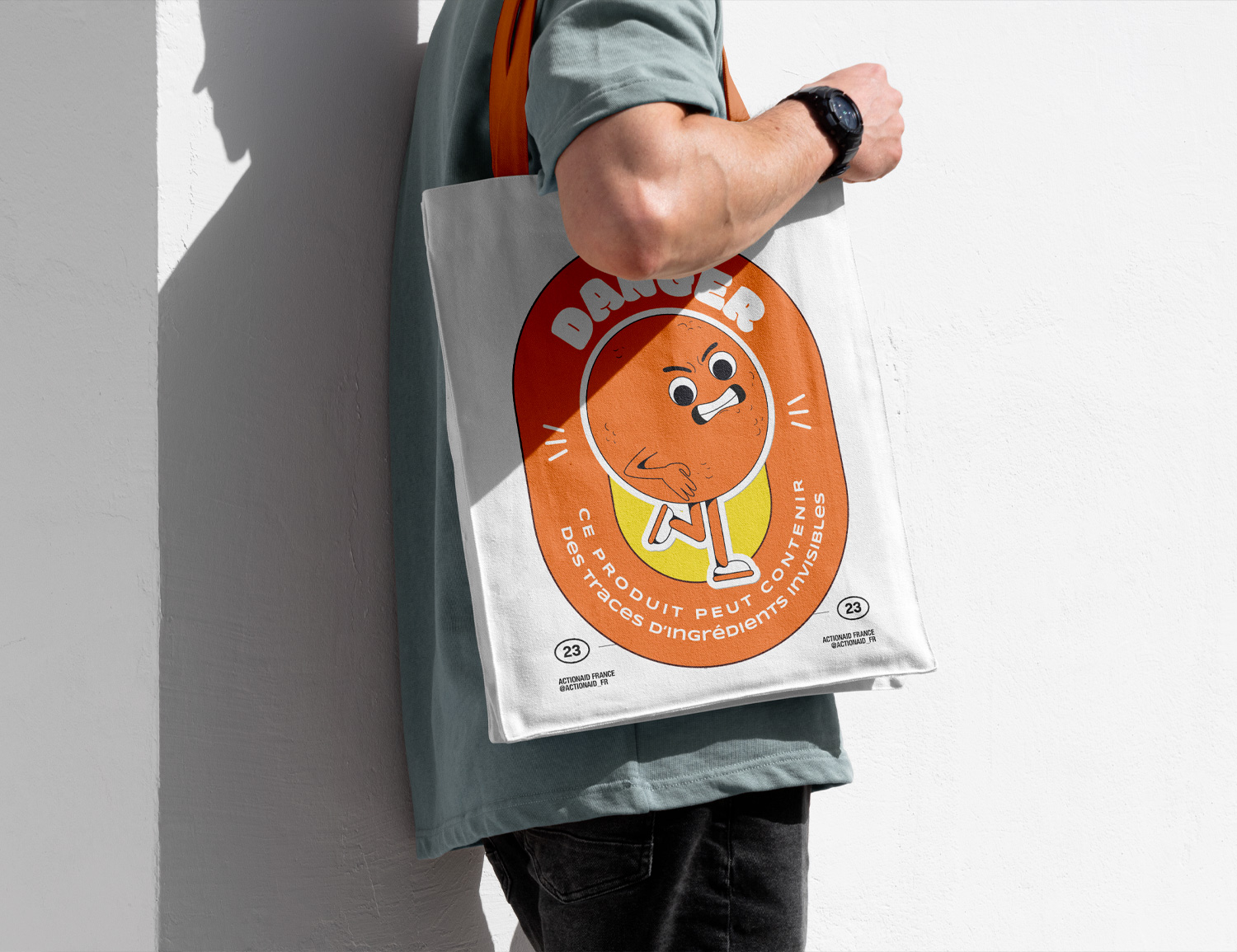 Homme portant un tote bag blanc avec un motif orange et un message 'DANGER : Ce produit peut contenir des traces d'ingrédients invisibles', illustrant une campagne de sensibilisation de @actionaid_fr.