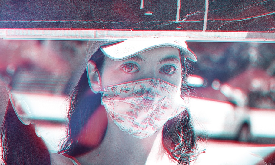 Photographie d'une jeune femme portant une visière et un masque facial à motifs floraux, tenant un panneau lors d'une manifestation ou d'un événement public. La photographie est prise à travers un prisme donnant une dimension supplémentaire à l'image, renforçant l'impact visuel de la scène capturée.