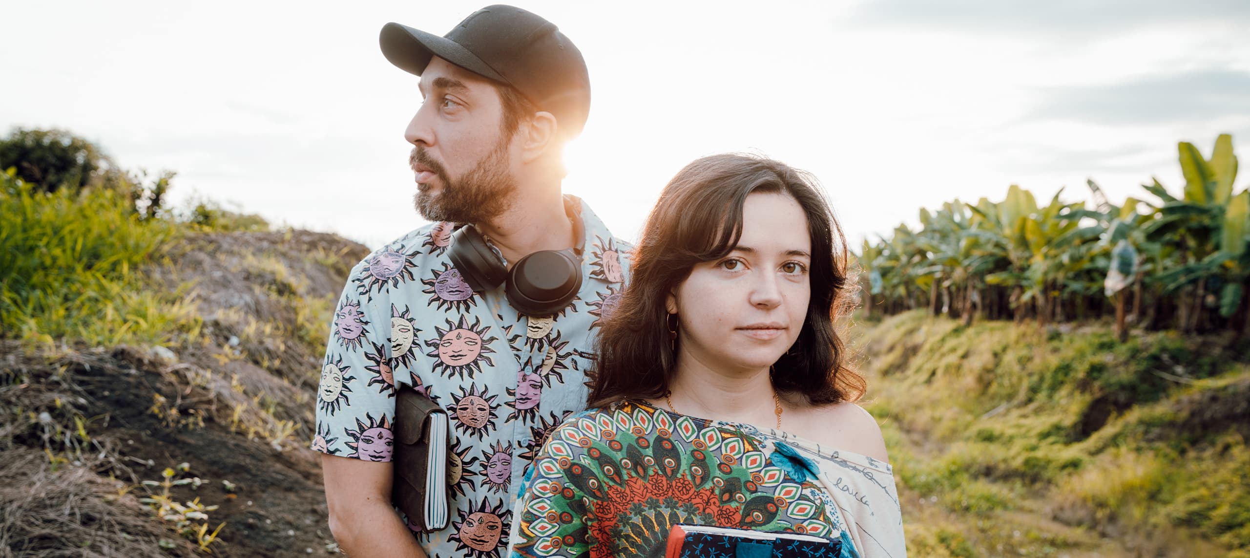 Portrait du rappeur Hippocampe Fou et de l'auteure de bande dessinée Blanche Sabbah devant une bananeraie verdoyante, lui portant une chemise à motifs de soleils et des écouteurs autour du cou, elle tenant un éventail coloré et un livre, avec le soleil se couchant en arrière-plan.