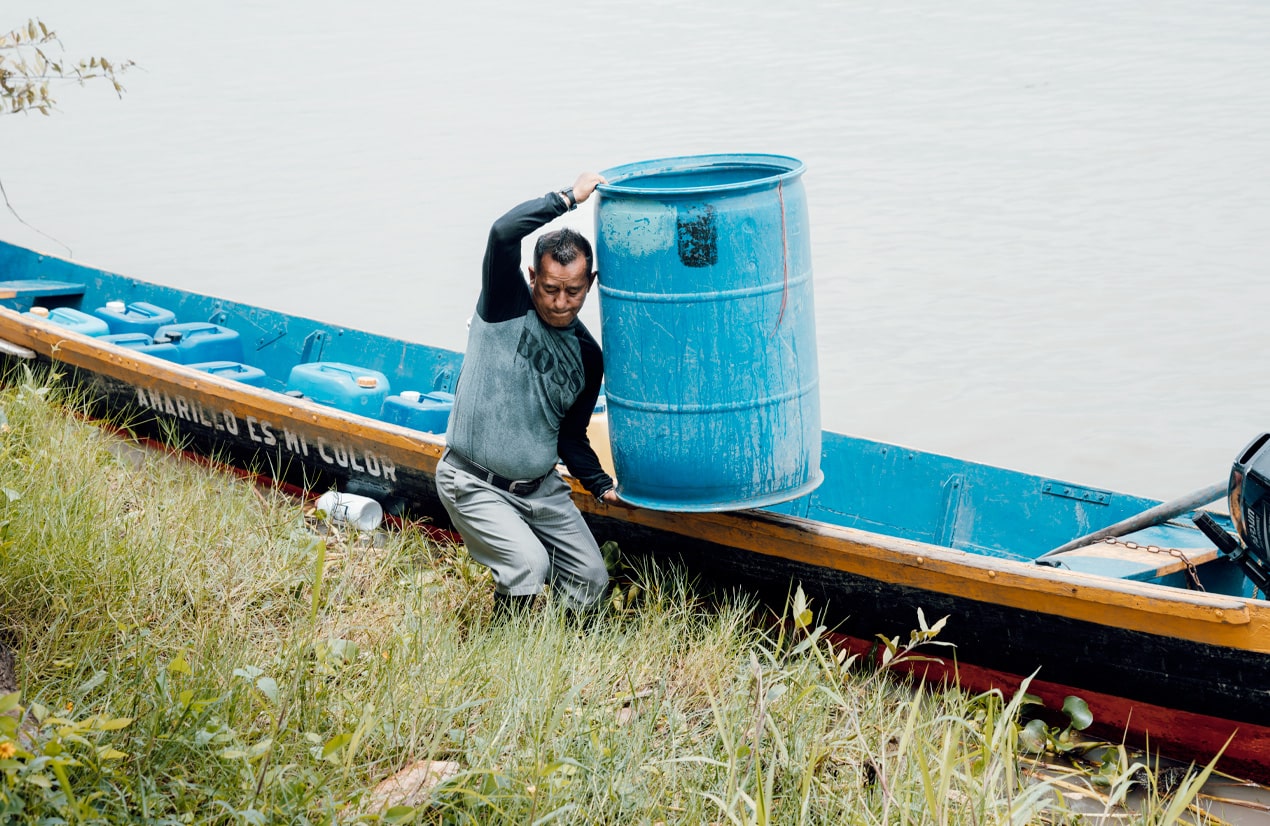 Un homme portant un pull gris et un pantalon, soulève un grand baril bleu depuis une barque en bois bleu et jaune sur la rive d'une rivière. La barque est remplie de bidons bleus et est ancrée près d'une zone herbeuse.