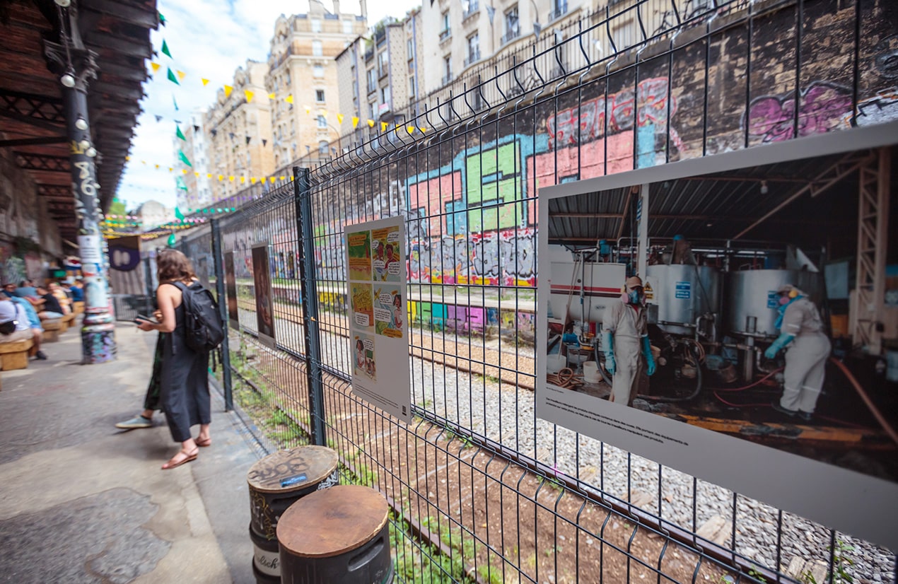 Une allée urbaine accueille une exposition photo en plein air, avec des images de grand format fixées sur un grillage. Les photos montrent des scènes variées, dont certaines semblent liées à la nutrition et à l'agriculture. Une femme se tient à distance, examinant les œuvres. L'allée est bordée de murs couverts de graffitis colorés et ornée de guirlandes de drapeaux, créant un environnement dynamique et artistique pour l'exposition.