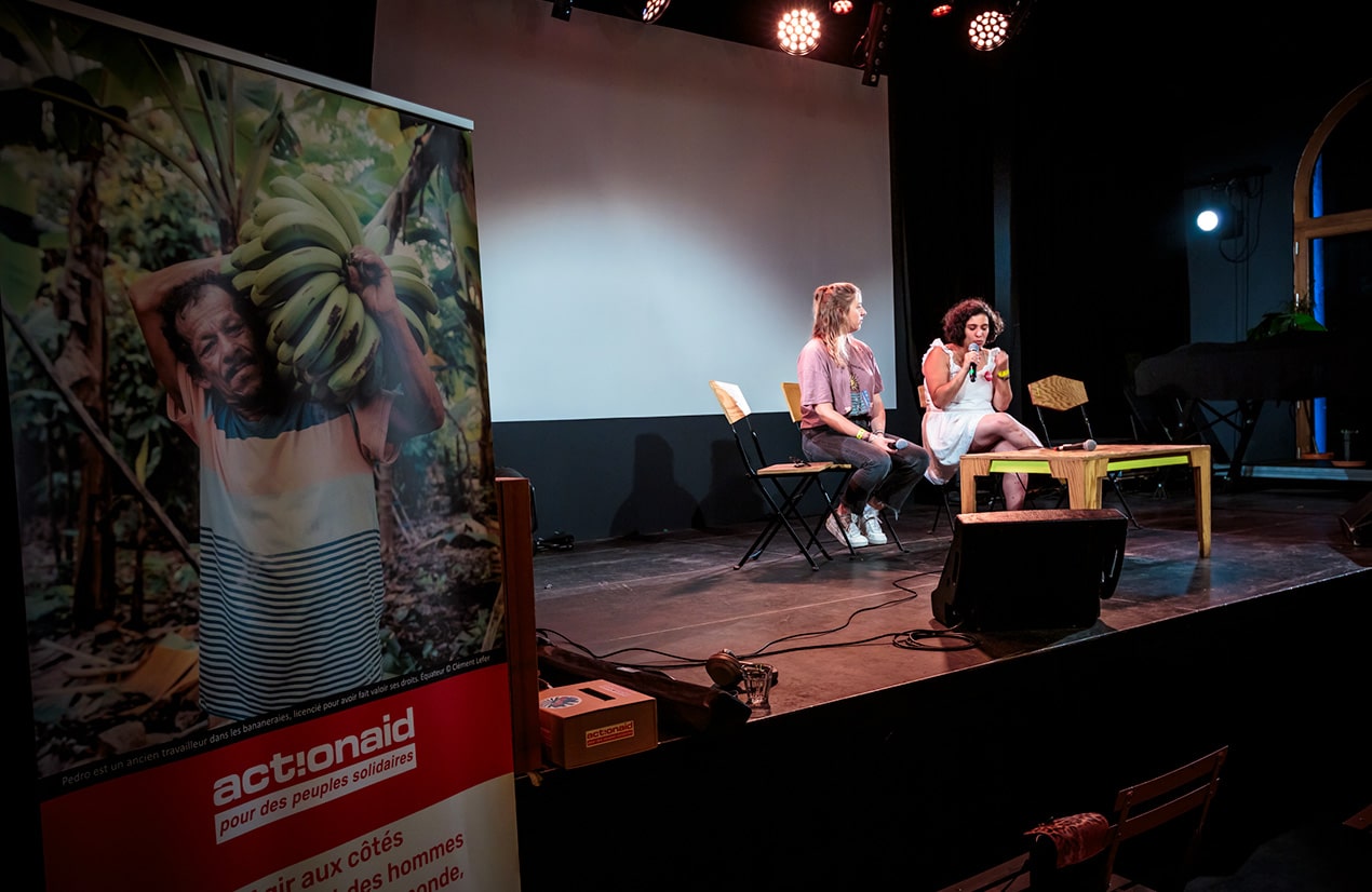 Deux femmes assises sur une scène lors d'un événement organisé par ActionAid. L'une porte une robe blanche et tient un micro, tandis que l'autre porte un t-shirt rose et un jean. À gauche, une bannière de l'organisation montre un ancien travailleur des bananeraies portant un régime de bananes. L'arrière-plan est sombre, avec des projecteurs éclairant la scène.