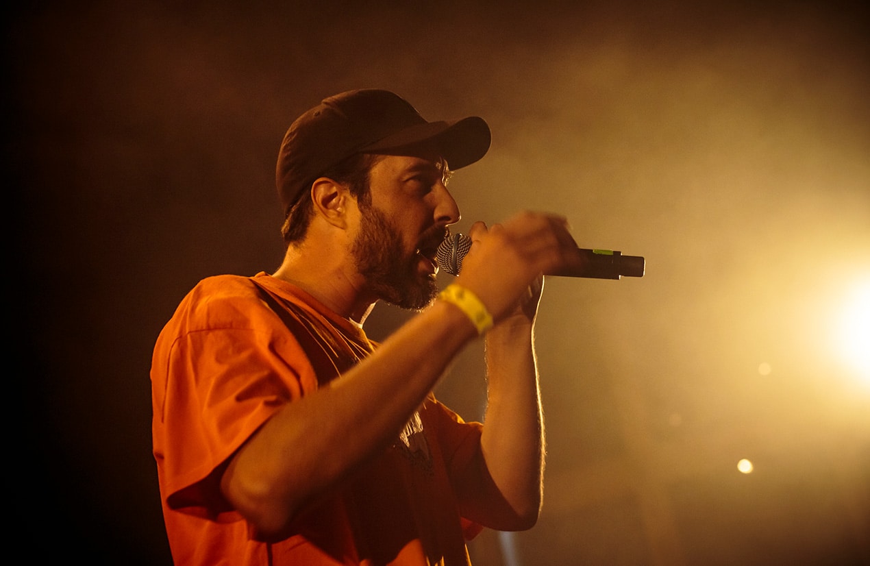 Photo du rappeur Hippocampe Fou, portant une casquette noire et un t-shirt orange, chantant dans un micro sur scène. L'éclairage de la scène est chaleureux et intense, créant une ambiance dramatique. L'arrière-plan est sombre, mettant en valeur l'artiste.