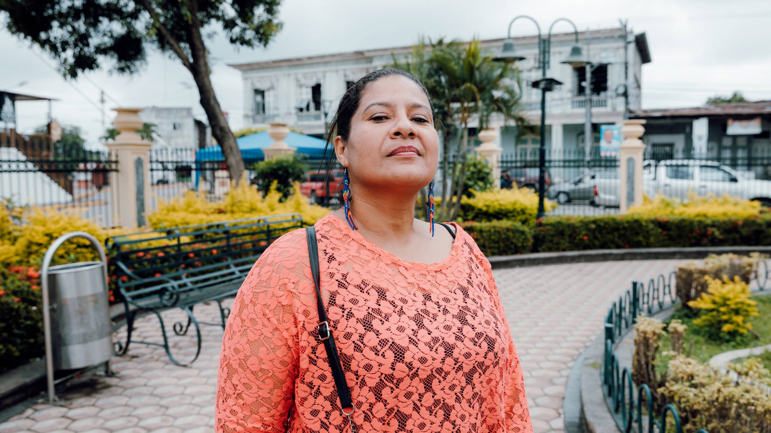 Portrait de Maricela, portant un haut orange en dentelle, debout dans un parc avec des bancs, des arbustes et des bâtiments en arrière-plan. Elle porte des boucles d'oreilles colorées et regarde fièrement l'objectif.