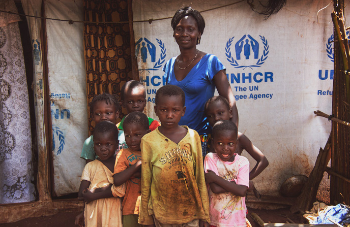 Femme centrafricaine debout avec un groupe d'enfants devant une tente marquée du logo de l'UNHCR dans un camp de réfugiés, illustrant le soutien et la protection des populations déplacées.