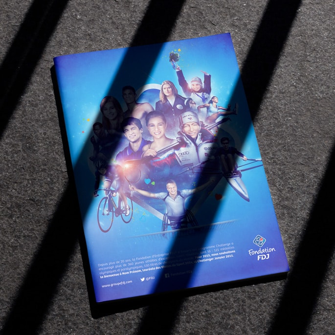 Brochure promotionnelle de la Fondation FDJ posée sur un sol bétonné avec ombres, illustrant divers athlètes en action dans un montage vibrant sur fond bleu, promouvant l'engagement sportif et le développement personnel.