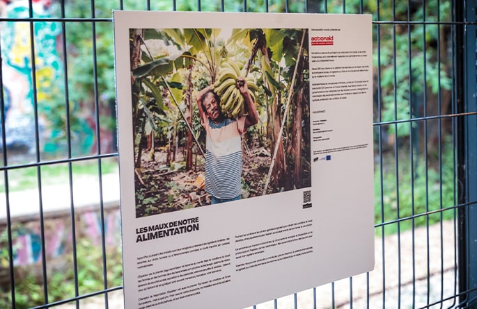 Panneau d'affichage suspendu sur une grille, montrant une photo d'une personne souriante tenant des bananes fraîchement récoltées sous des plants de bananiers. Le titre en haut du panneau indique 'LES MAUX DE NOTRE ALIMENTATION', en référence à l'exposition éponyme. En dessous de la photo, un texte descriptif accompagne l'image. En bas, le logo d'ActionAid est visible, ainsi qu'un code QR pour plus d'informations.